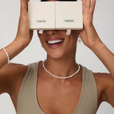 A smiling woman holds two beige VANCHI boxes over her eyes like glasses. She wears a taupe tank top, the Flora Multi Pearl Toggle Choker in Gold, along with a matching bracelet and pearl earrings against a plain background.
