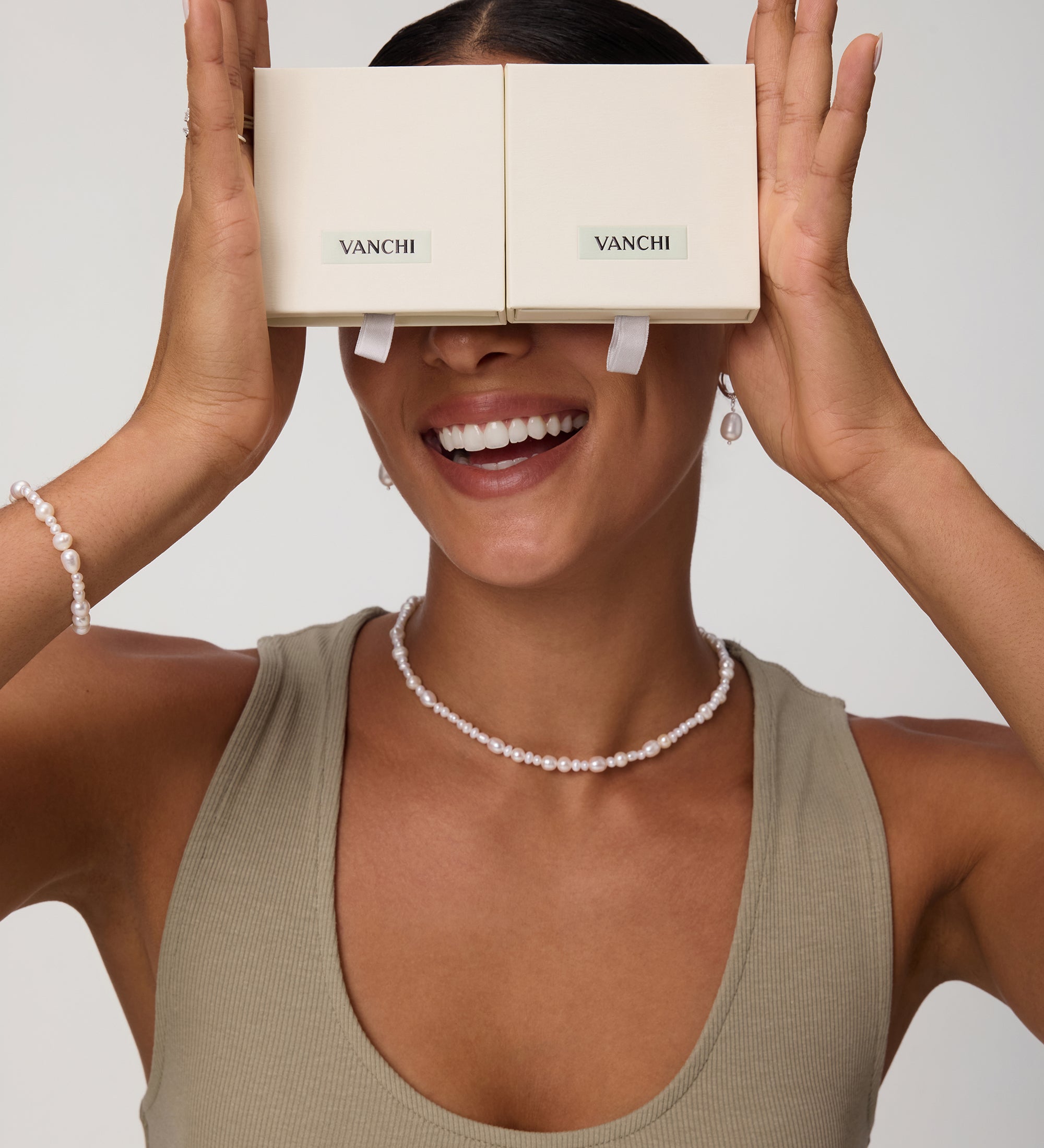 A smiling woman holds two beige VANCHI boxes over her eyes like glasses. She wears a taupe tank top, the Flora Multi Pearl Toggle Choker in Gold, along with a matching bracelet and pearl earrings against a plain background.