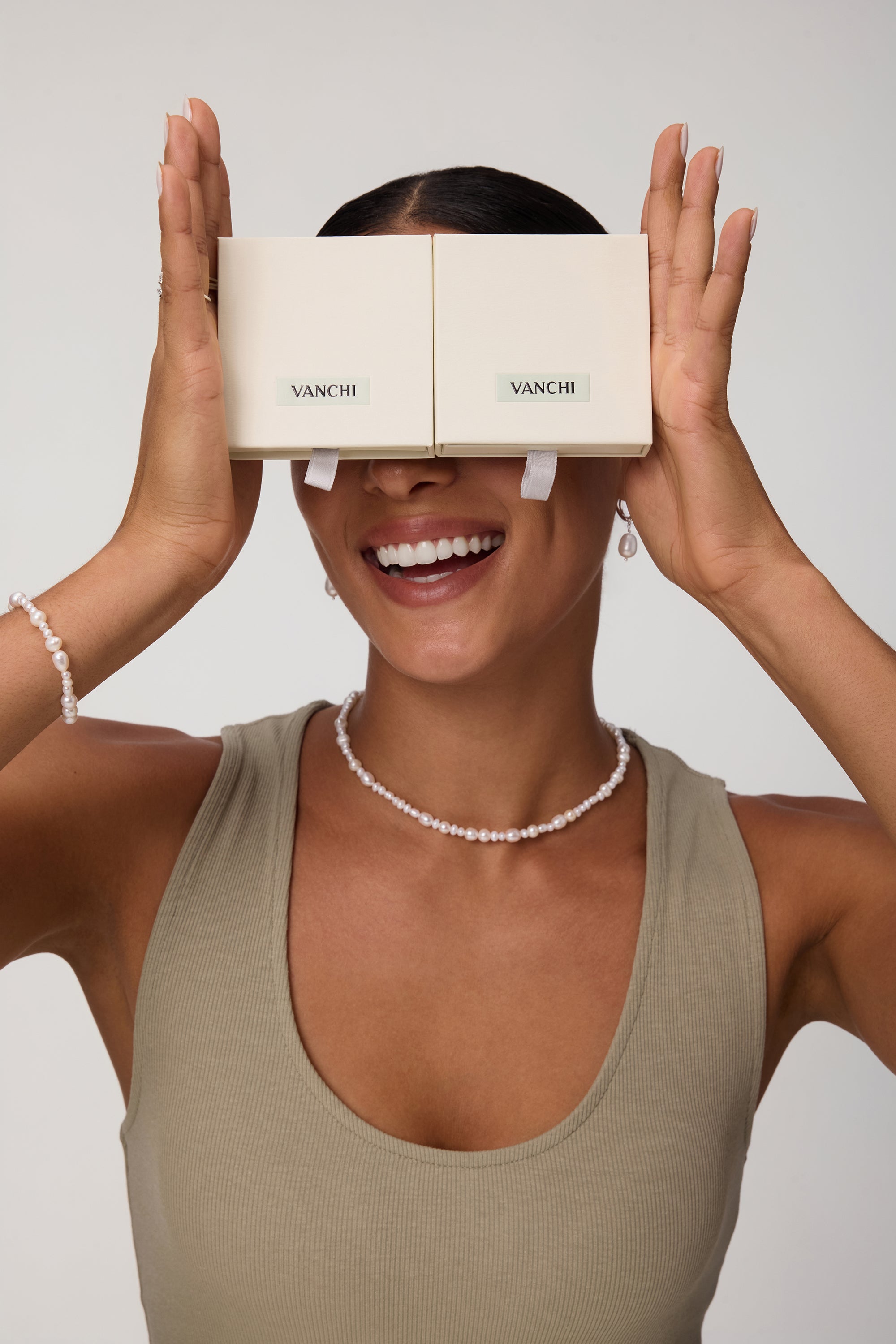 A smiling woman holds two beige VANCHI boxes over her eyes like glasses. She wears a taupe tank top, the Flora Multi Pearl Toggle Choker in Gold, along with a matching bracelet and pearl earrings against a plain background.
