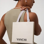 A woman wears the Freshwater Pearl Charm Huggies in Silver with a matching necklace, holding a white VANCHI shopping bag over her shoulder. She is dressed in a sleeveless olive dress and is facing slightly away from the camera.