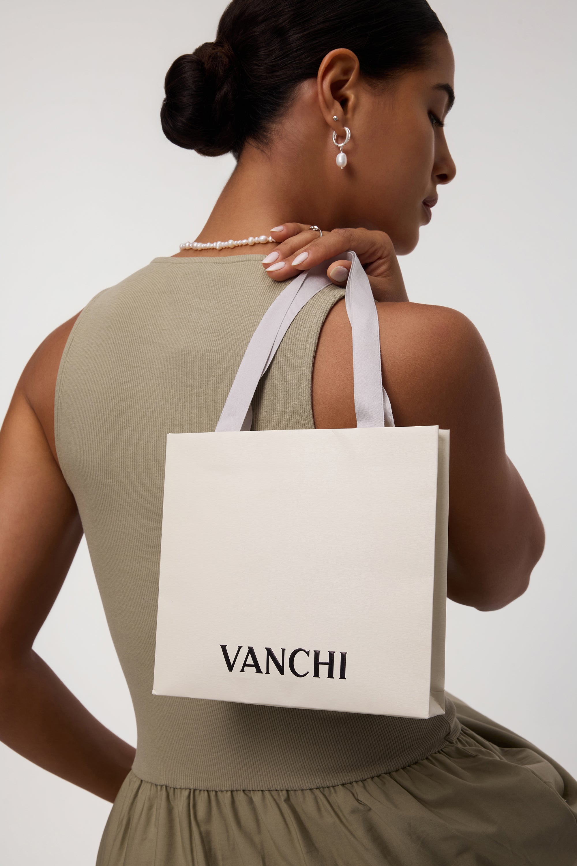 A woman wears the Freshwater Pearl Charm Huggies in Silver with a matching necklace, holding a white VANCHI shopping bag over her shoulder. She is dressed in a sleeveless olive dress and is facing slightly away from the camera.