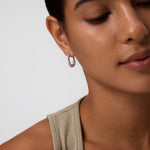 A close-up of a woman with smooth skin and dark hair pulled back, wearing a light beige sleeveless top and Nouf Pavé Hoop Earrings in Gold, set against a light background with a calm expression.
