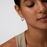 A close-up of a woman with smooth skin and dark hair pulled back, wearing a light beige sleeveless top and Nouf Pavé Hoop Earrings in Gold, set against a light background with a calm expression.