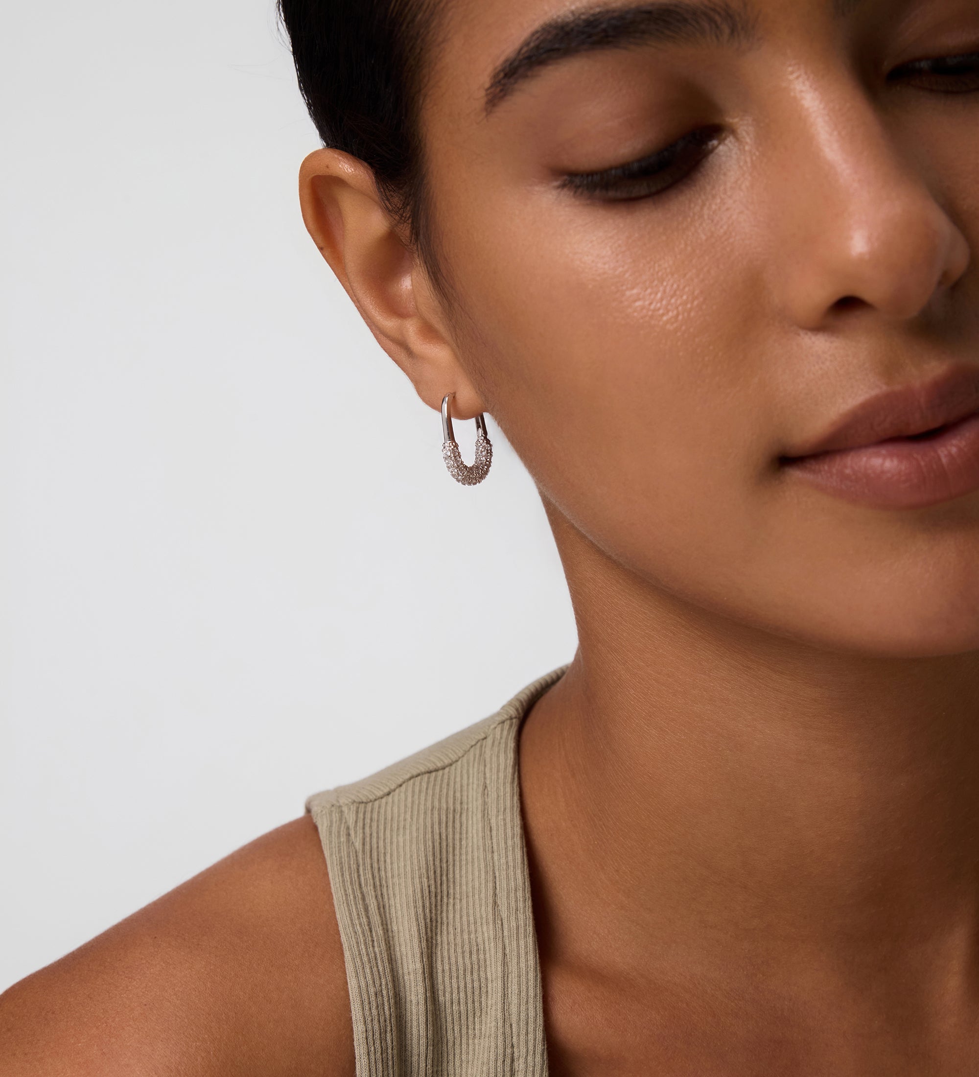 A close-up of a woman with smooth skin and dark hair pulled back, wearing a light beige sleeveless top and Nouf Pavé Hoop Earrings in Gold, set against a light background with a calm expression.