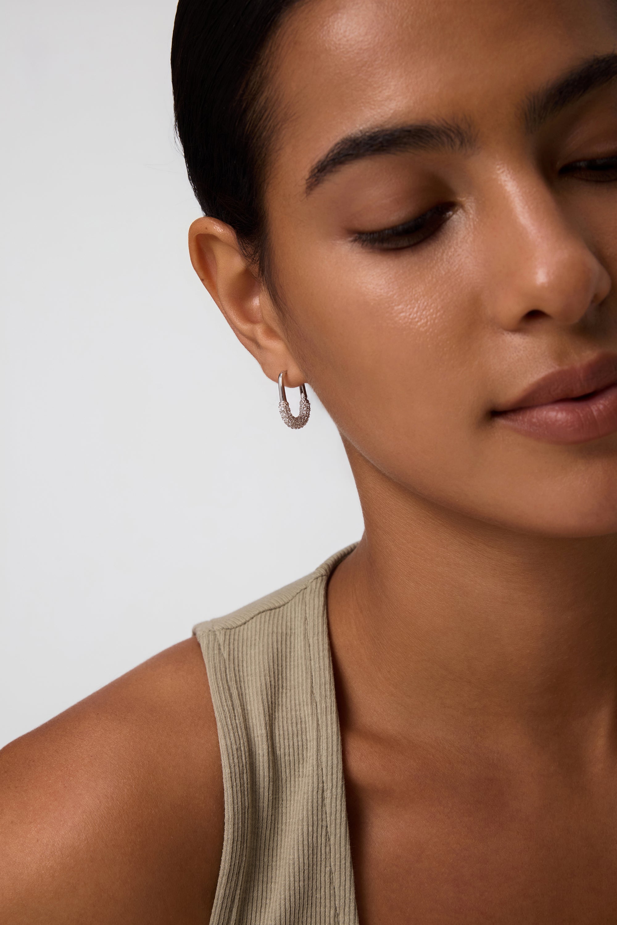 A close-up of a woman with smooth skin and dark hair pulled back, wearing a light beige sleeveless top and Nouf Pavé Hoop Earrings in Gold, set against a light background with a calm expression.