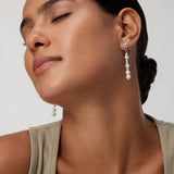 A woman with closed eyes and smooth skin wears a beige sleeveless top and the Knot Multi Pearl Drop Earrings in Gold. Her dark hair is styled in a low bun against a plain background.