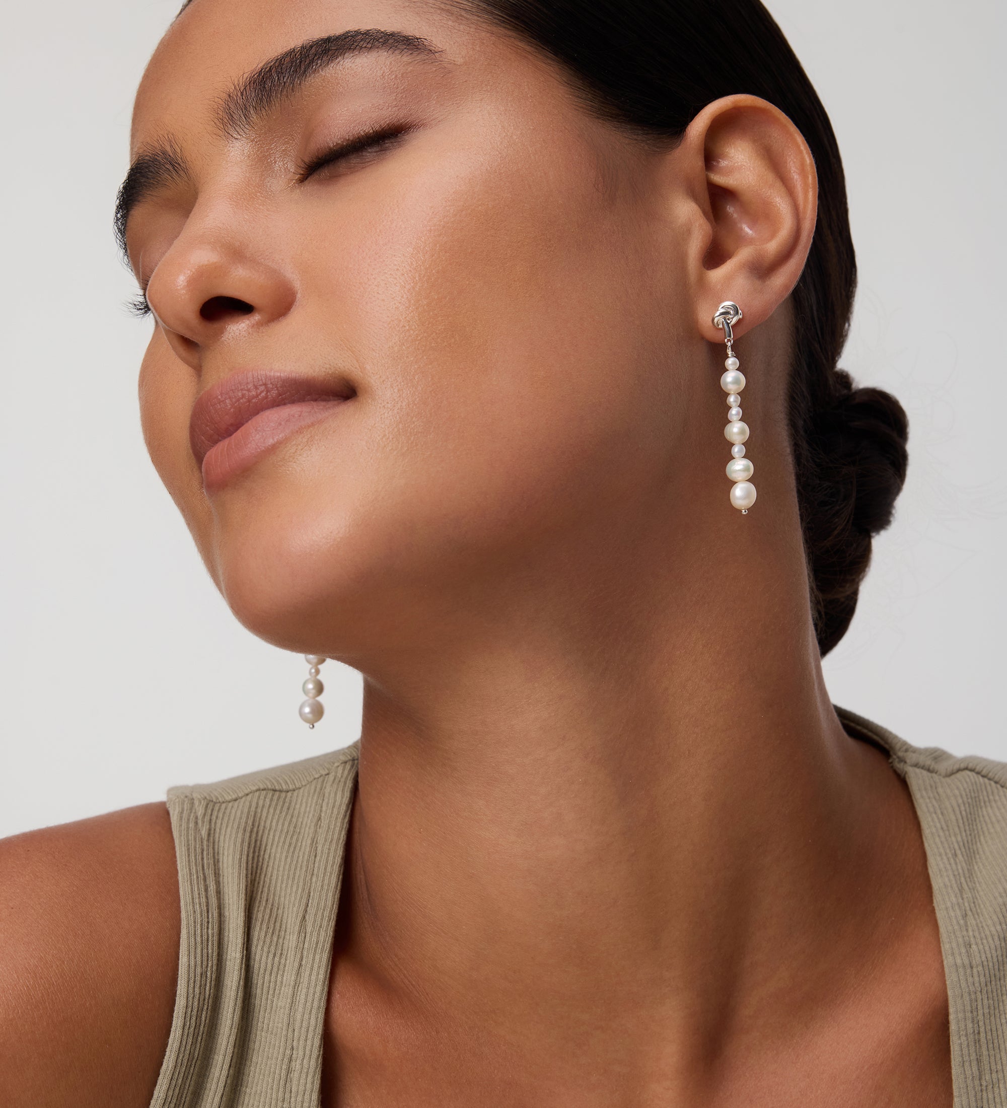 A woman with closed eyes and smooth skin wears a beige sleeveless top and the Knot Multi Pearl Drop Earrings in Gold. Her dark hair is styled in a low bun against a plain background.