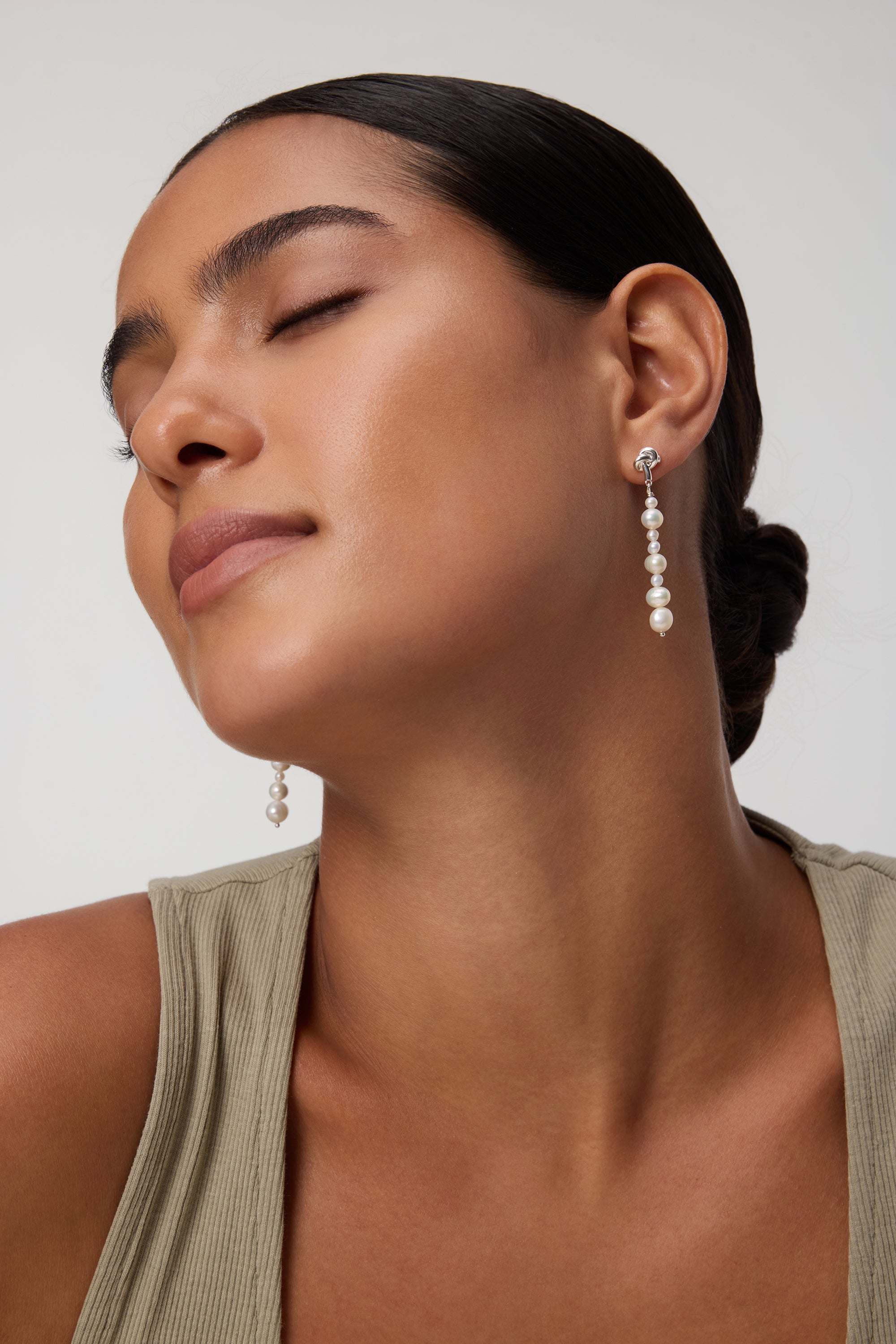 A woman with closed eyes and smooth skin wears a beige sleeveless top and the Knot Multi Pearl Drop Earrings in Gold. Her dark hair is styled in a low bun against a plain background.