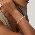 A woman rests her hand near her ear, wearing a sleeveless beige top and two bracelets: one adorned with pearls and the other—the Rhea Molten Cascade Bracelet in Gold—featuring striking gold plating, small dangling charms, and a subtle hammered texture. The background is light gray.