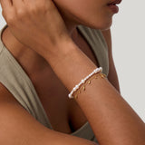 A woman rests her hand near her ear, wearing a sleeveless beige top and two bracelets: one adorned with pearls and the other—the Rhea Molten Cascade Bracelet in Gold—featuring striking gold plating, small dangling charms, and a subtle hammered texture. The background is light gray.