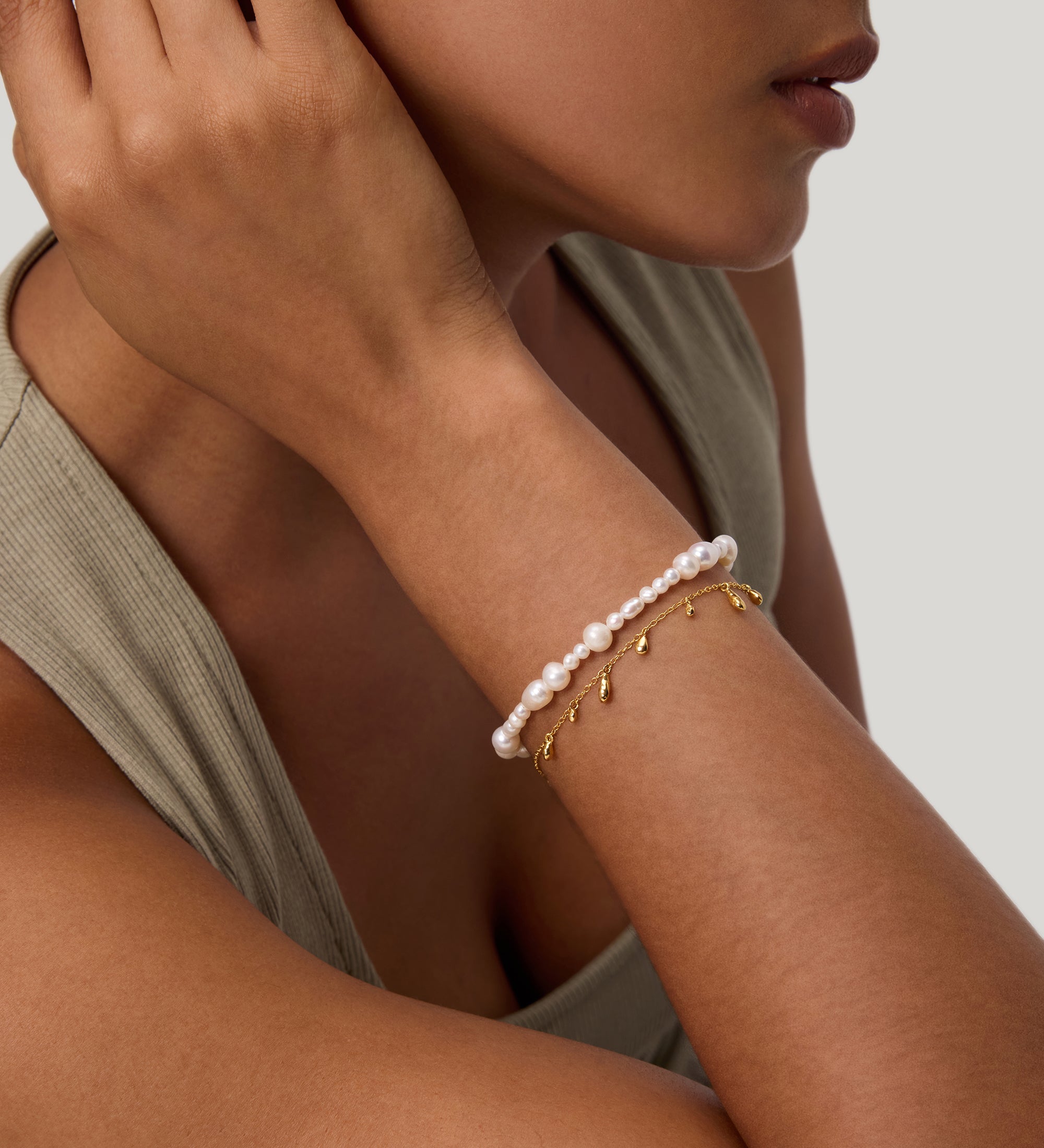 A woman rests her hand near her ear, wearing a sleeveless beige top and two bracelets: one adorned with pearls and the other—the Rhea Molten Cascade Bracelet in Gold—featuring striking gold plating, small dangling charms, and a subtle hammered texture. The background is light gray.