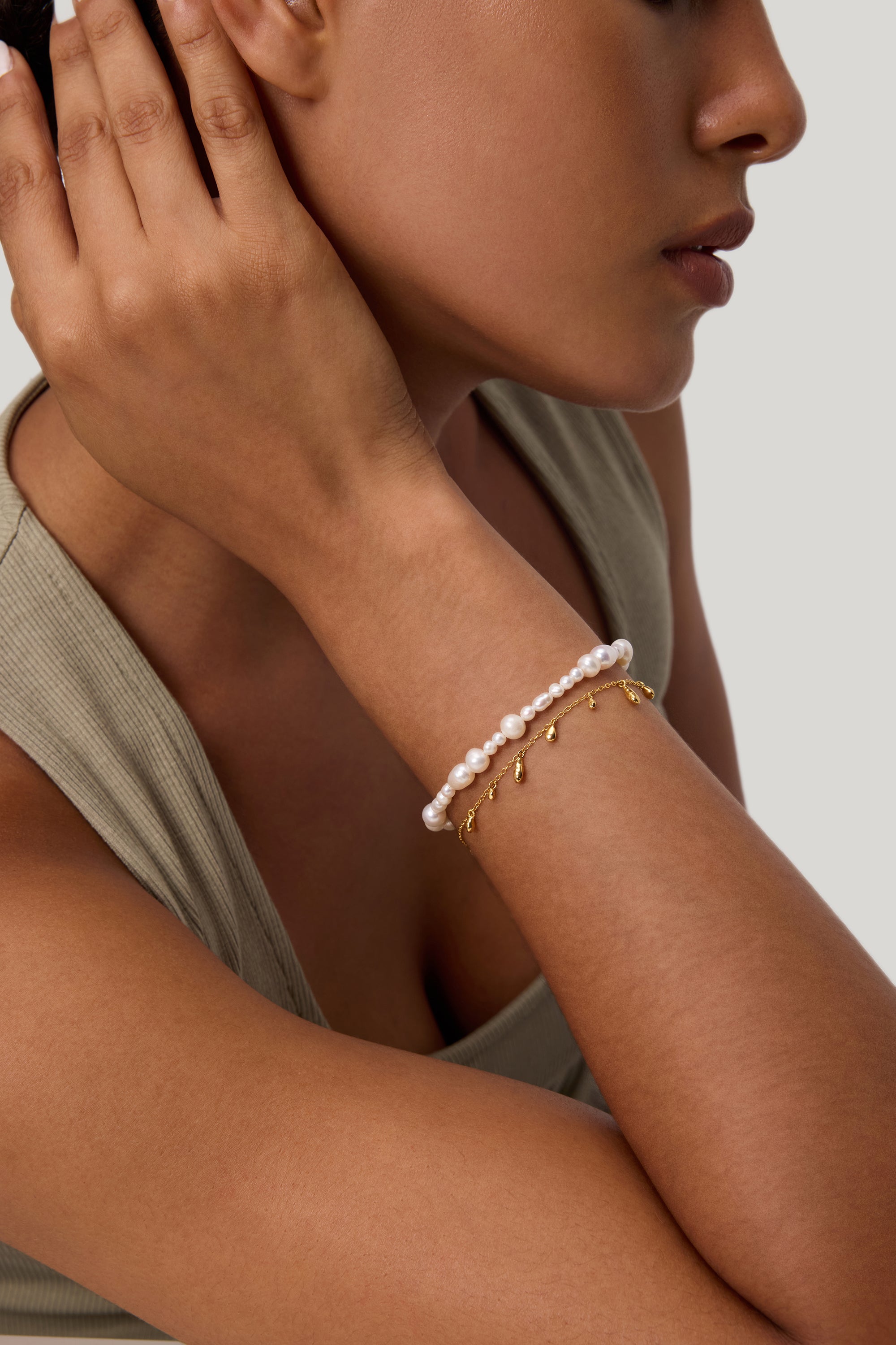 A woman rests her hand near her ear, wearing a sleeveless beige top and two bracelets: one adorned with pearls and the other—the Rhea Molten Cascade Bracelet in Gold—featuring striking gold plating, small dangling charms, and a subtle hammered texture. The background is light gray.