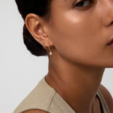 A woman with smooth skin and dark hair pulled back wears a sleeveless beige top and three gold earrings, including the Flora Crystal Charm Huggies in Gold. The image features the right side of her face and ear.