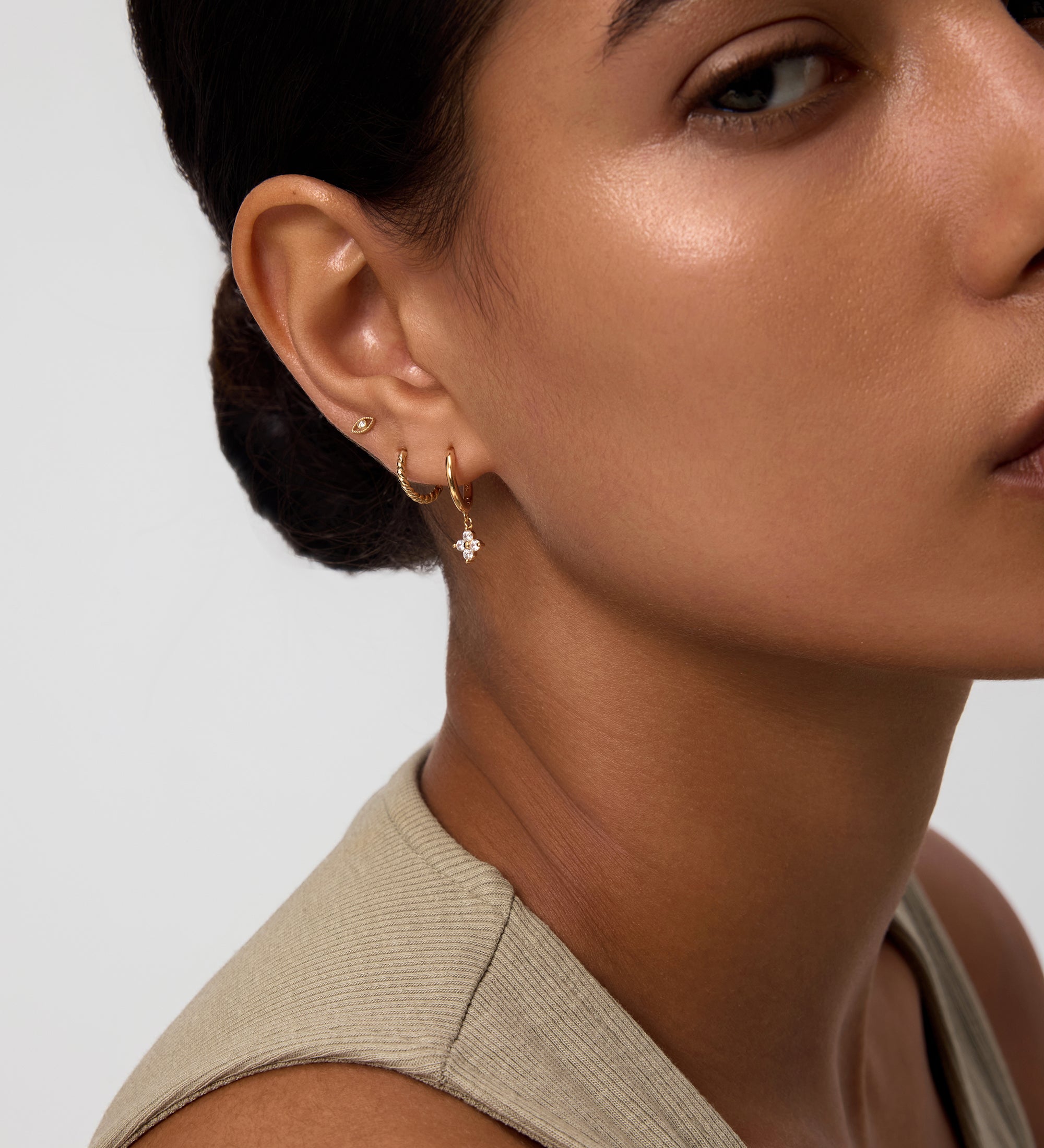 A woman with smooth skin and dark hair pulled back wears a sleeveless beige top and three gold earrings, including the Flora Crystal Charm Huggies in Gold. The image features the right side of her face and ear.