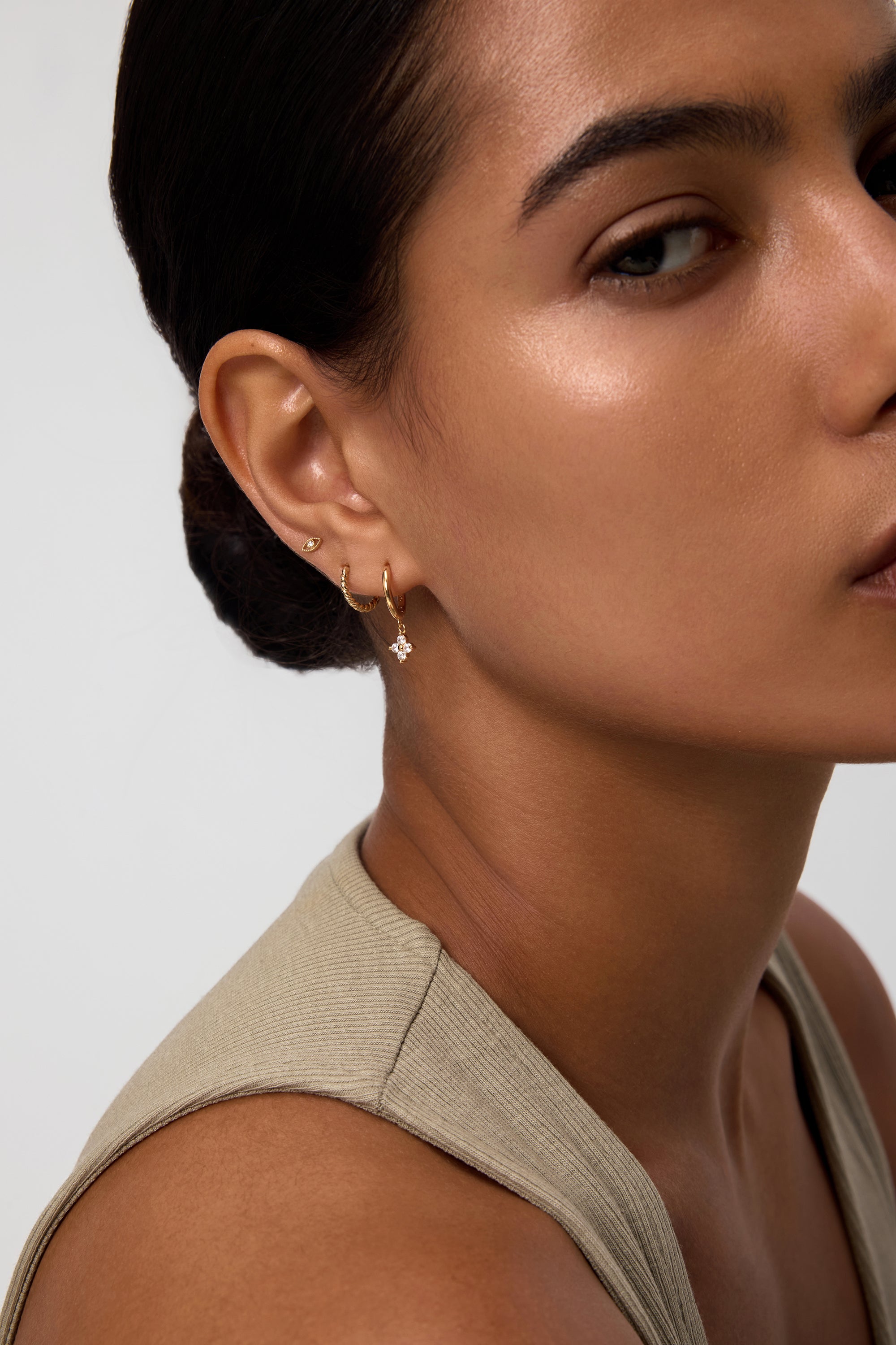 A woman with smooth skin and dark hair pulled back wears a sleeveless beige top and three gold earrings, including the Flora Crystal Charm Huggies in Gold. The image features the right side of her face and ear.