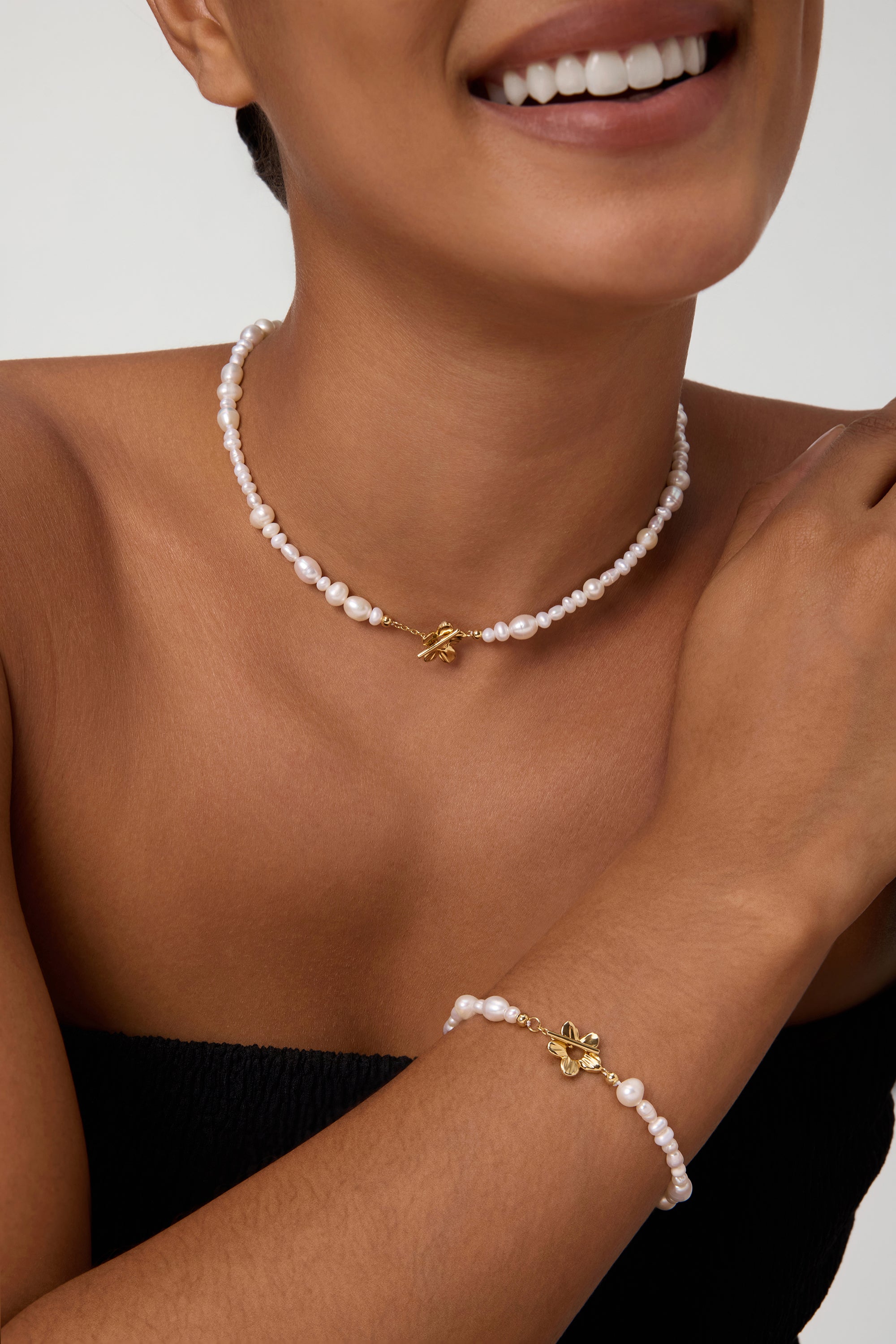Wearing the Flora Multi Pearl Toggle Choker in Gold, a smiling person showcases its elegant freshwater pearls, delicate gold charm, and sophisticated floral toggle clasp against a plain background.