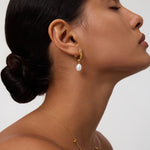 A woman with smooth skin and dark hair in a low bun wears the Freshwater Pearl Charm Huggies in Gold, their delicate pearls dangling from gold-plated hoops. She is shown in profile with her eyes closed against a plain background—an effortless, everyday essential look.