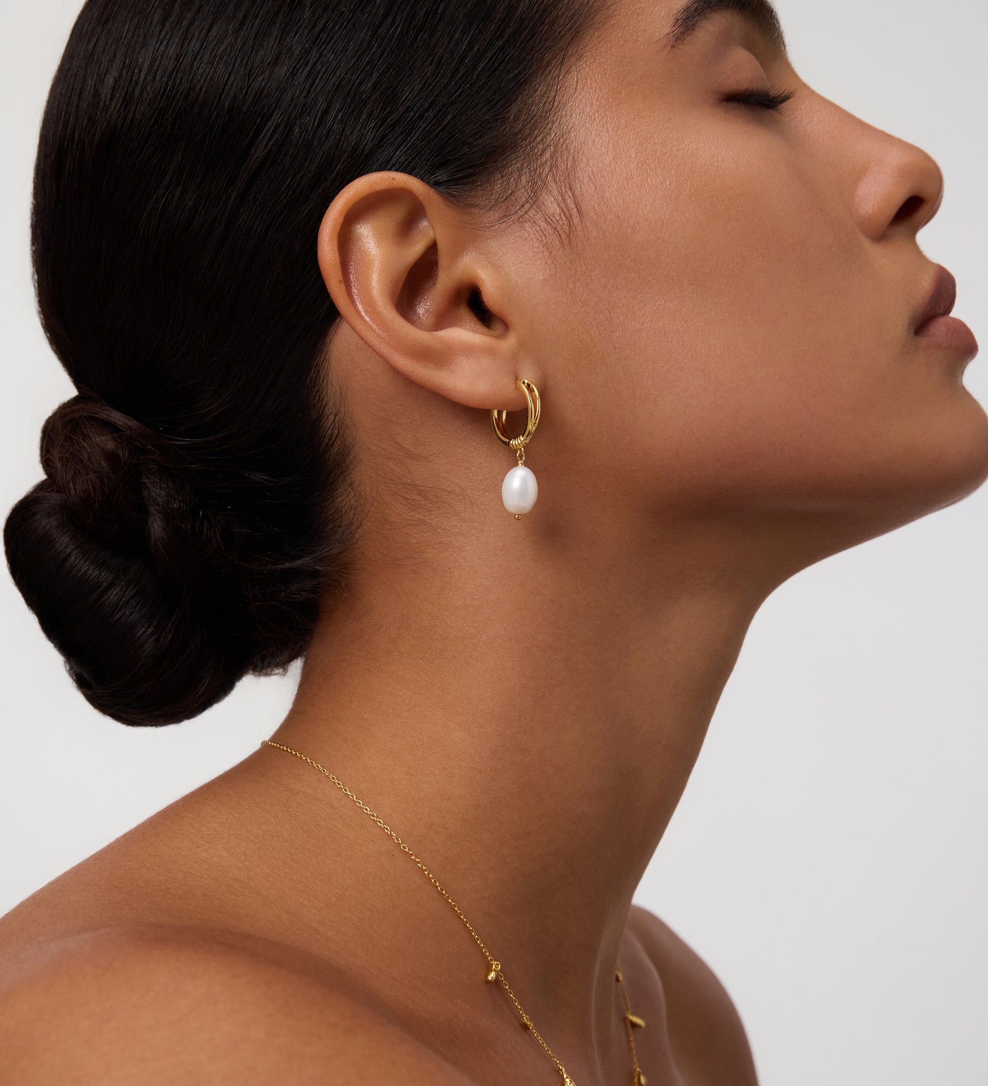 A woman with smooth skin and dark hair styled in a low bun wears minimalist jewelry, featuring the Freshwater Pearl Charm Hoops in Gold and a delicate gold necklace. She faces sideways with her eyes closed against a plain background.