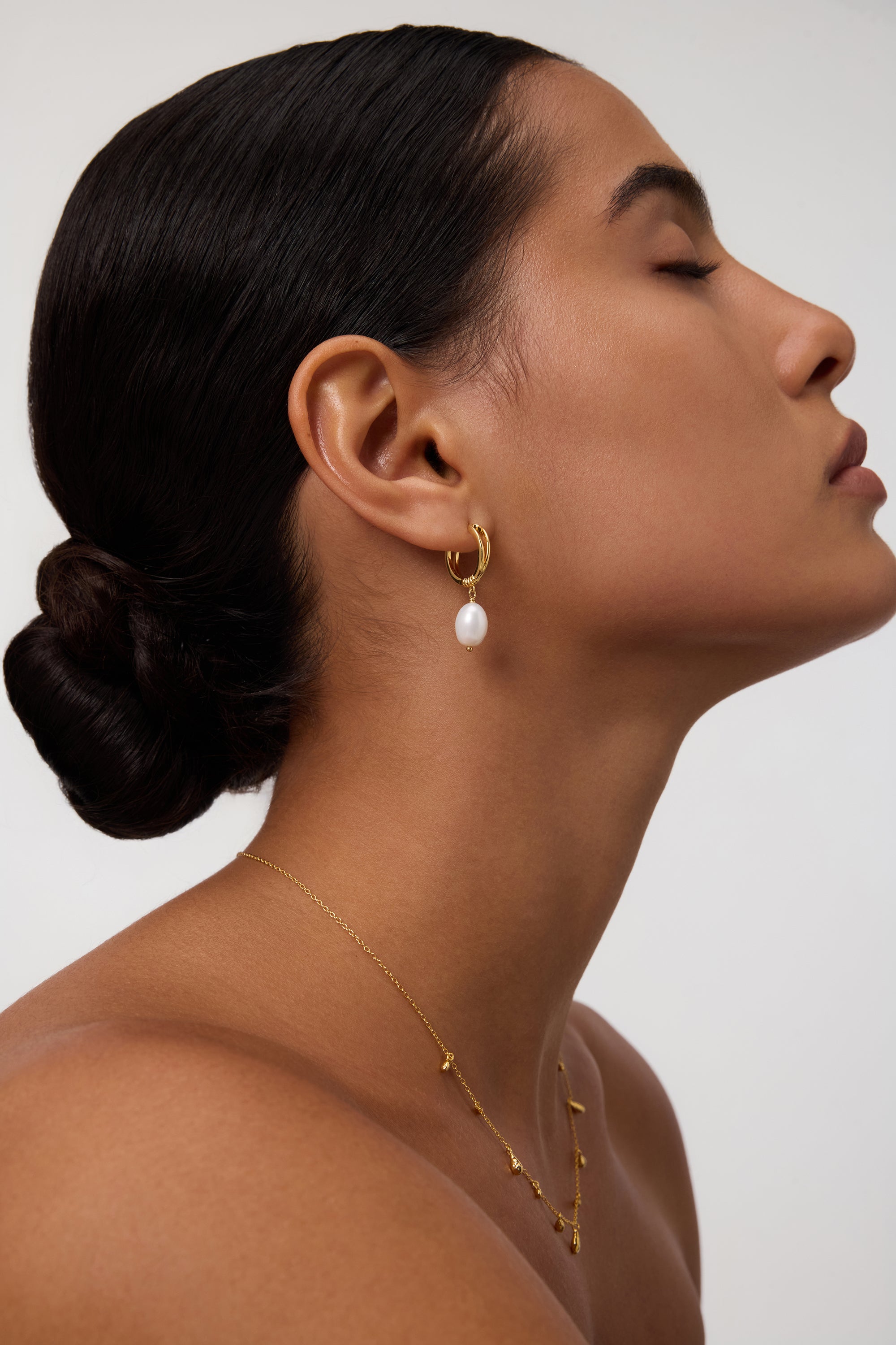 A woman with smooth skin and dark hair in a low bun wears the Freshwater Pearl Charm Huggies in Gold, their delicate pearls dangling from gold-plated hoops. She is shown in profile with her eyes closed against a plain background—an effortless, everyday essential look.