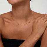 A woman, smiling with one hand resting on her shoulder against a plain background, wears a strapless black top, a gold drop necklace, a matching bracelet, and the Freshwater Pearl Charm Huggies in Gold—an everyday essential.