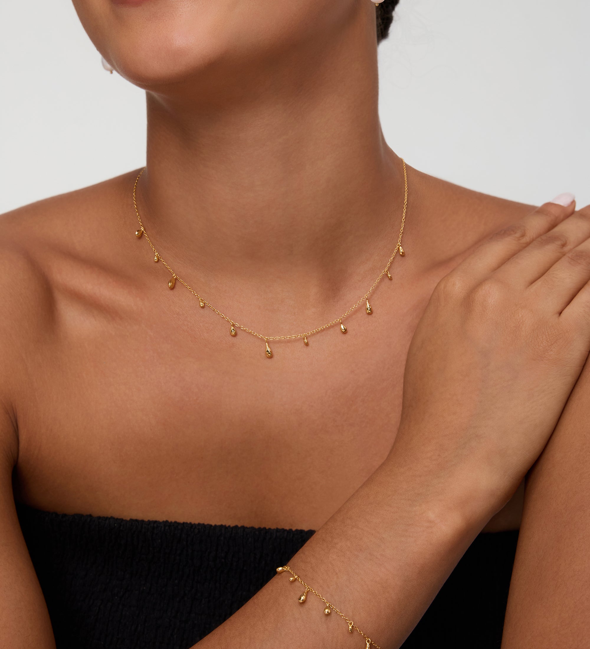 A woman, smiling with one hand resting on her shoulder against a plain background, wears a strapless black top, a gold drop necklace, a matching bracelet, and the Freshwater Pearl Charm Huggies in Gold—an everyday essential.