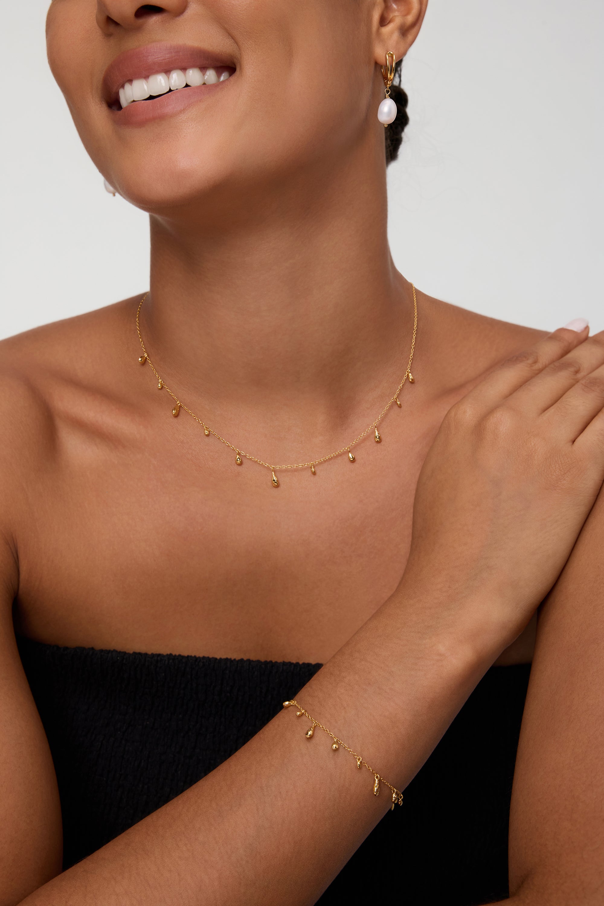 A woman, smiling with one hand resting on her shoulder against a plain background, wears a strapless black top, a gold drop necklace, a matching bracelet, and the Freshwater Pearl Charm Huggies in Gold—an everyday essential.