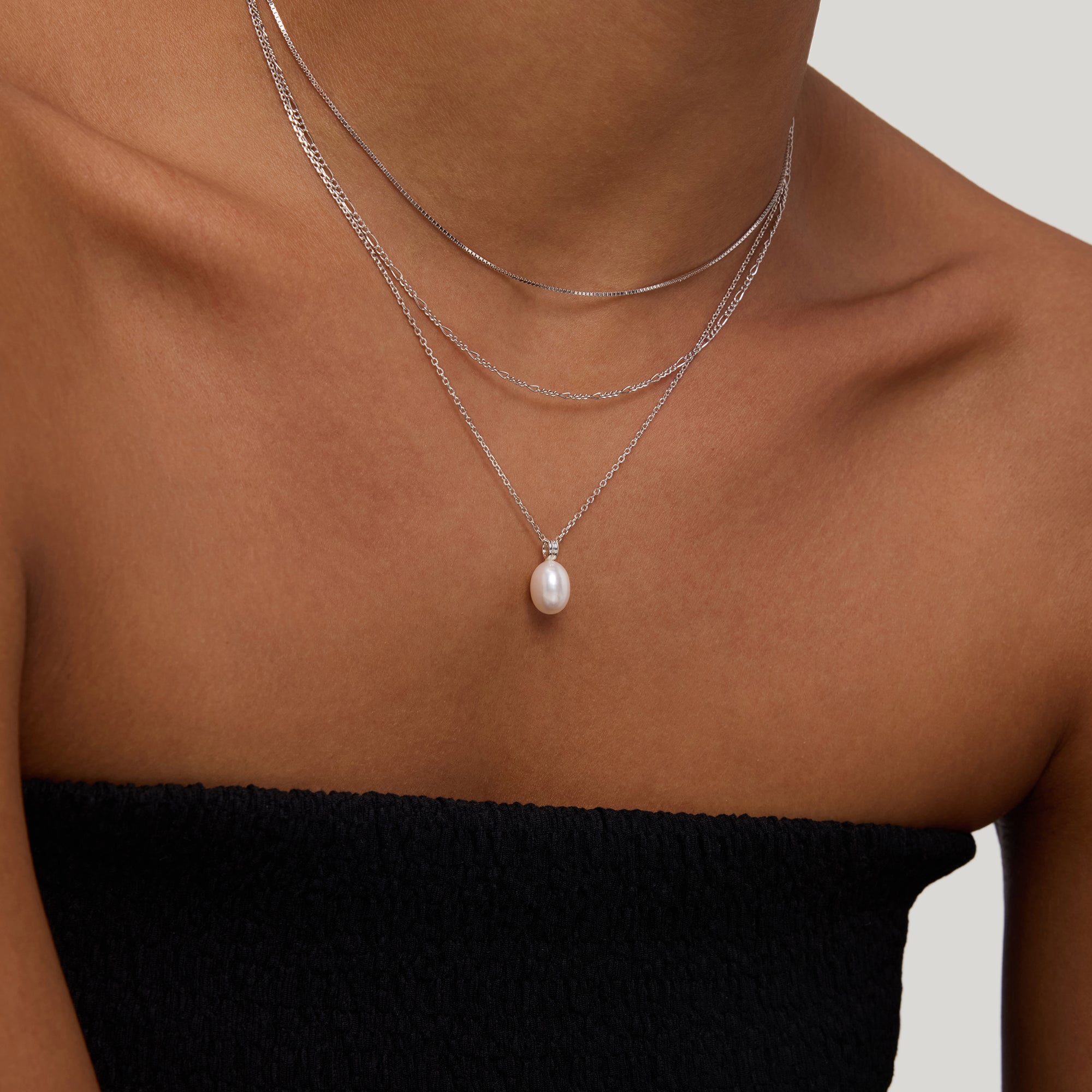 A woman models the Figaro And Box Chain Layered Necklace in Silver—three delicate sterling silver chains, including one with a pearl pendant—over a black strapless top against a neutral background.