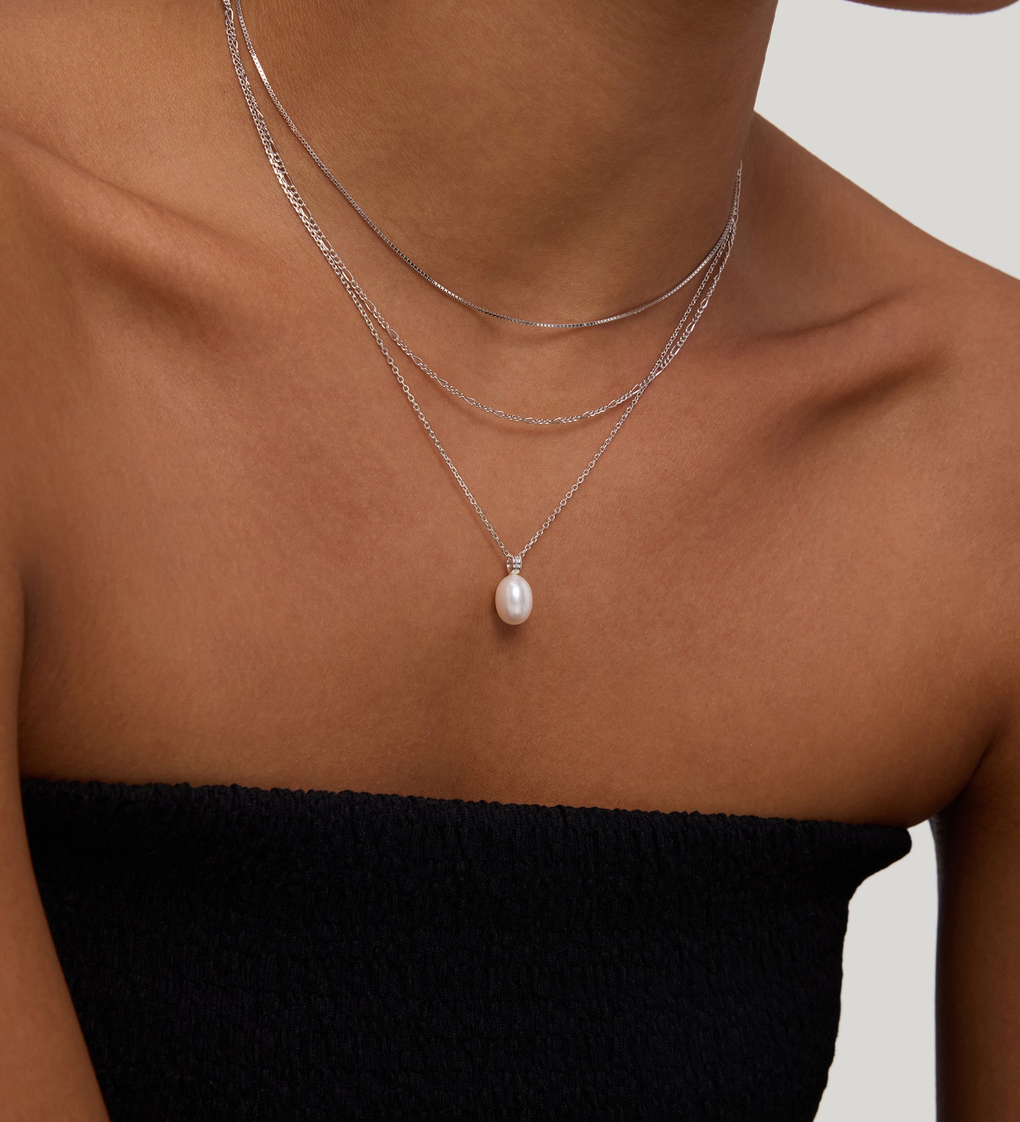 A woman models the Figaro And Box Chain Layered Necklace in Silver—three delicate sterling silver chains, including one with a pearl pendant—over a black strapless top against a neutral background.
