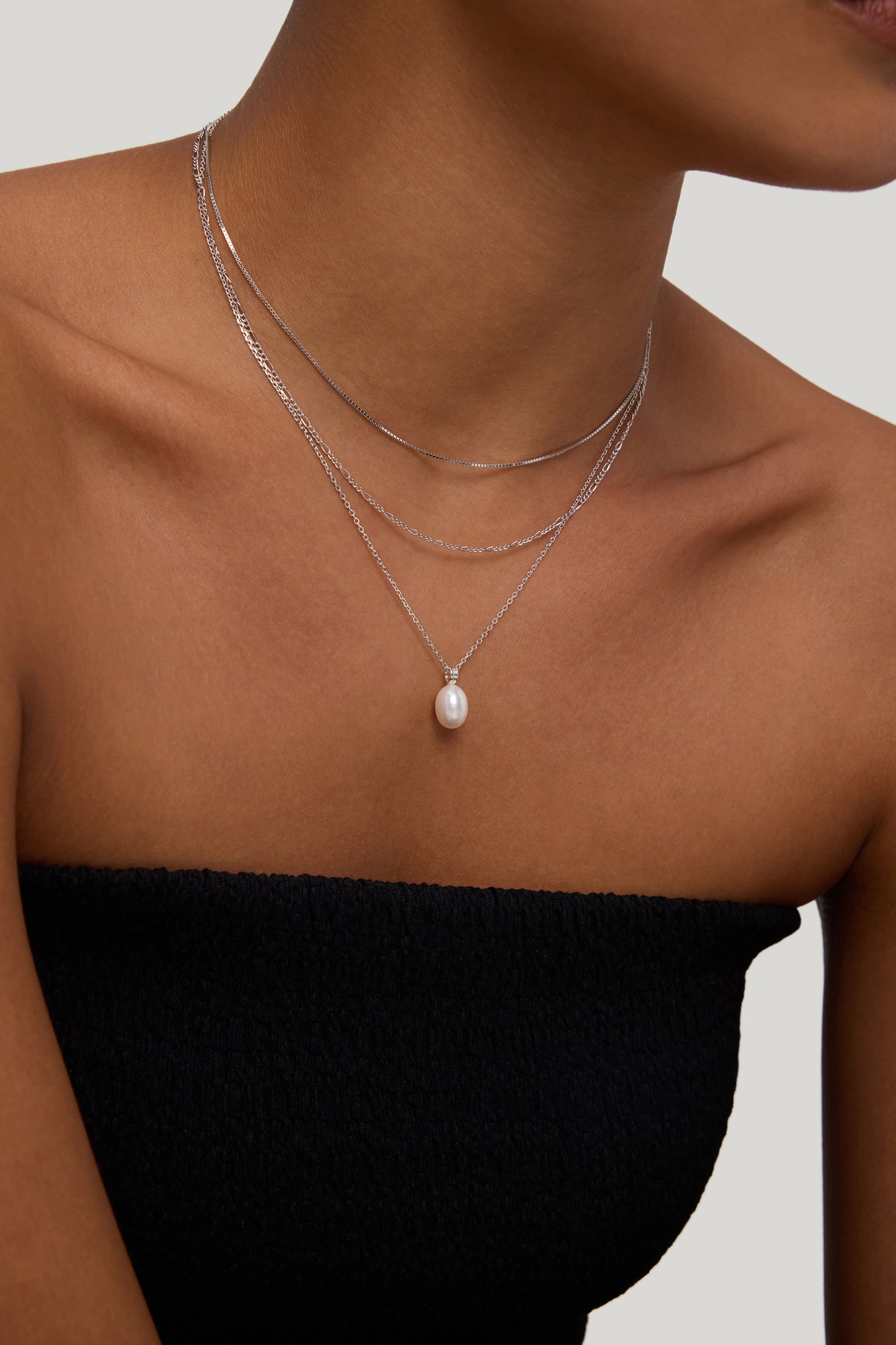 A woman models the Figaro And Box Chain Layered Necklace in Silver—three delicate sterling silver chains, including one with a pearl pendant—over a black strapless top against a neutral background.