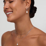 A woman with smooth, dark hair in a low bun smiles with her eyes closed, wearing a strapless black top and the Figaro And Box Chain Layered Necklace in Silver with a pearl pendant, paired with matching pearl drop earrings against a neutral background.