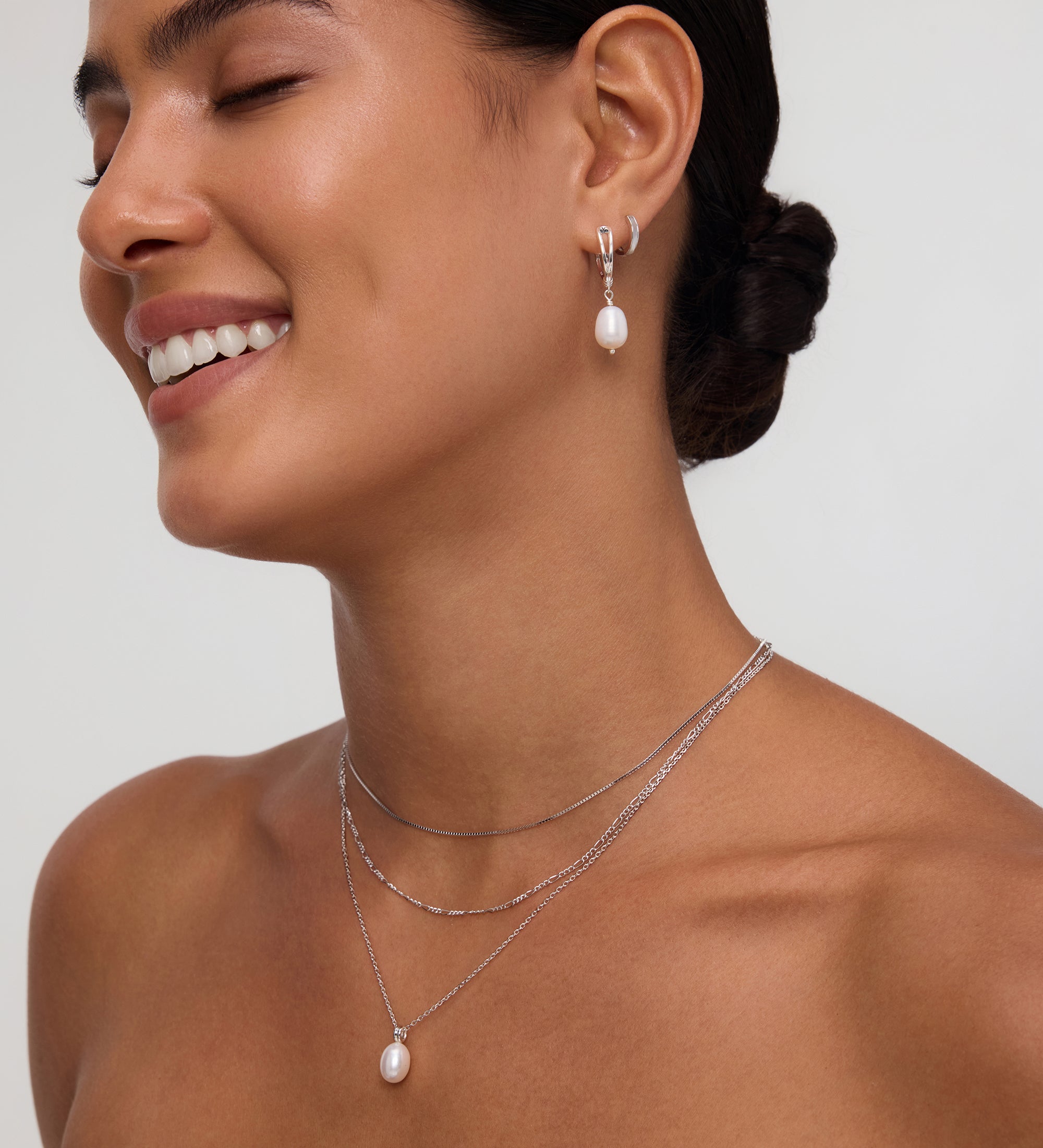 A woman with smooth, dark hair in a low bun smiles with her eyes closed, wearing a strapless black top and the Figaro And Box Chain Layered Necklace in Silver with a pearl pendant, paired with matching pearl drop earrings against a neutral background.