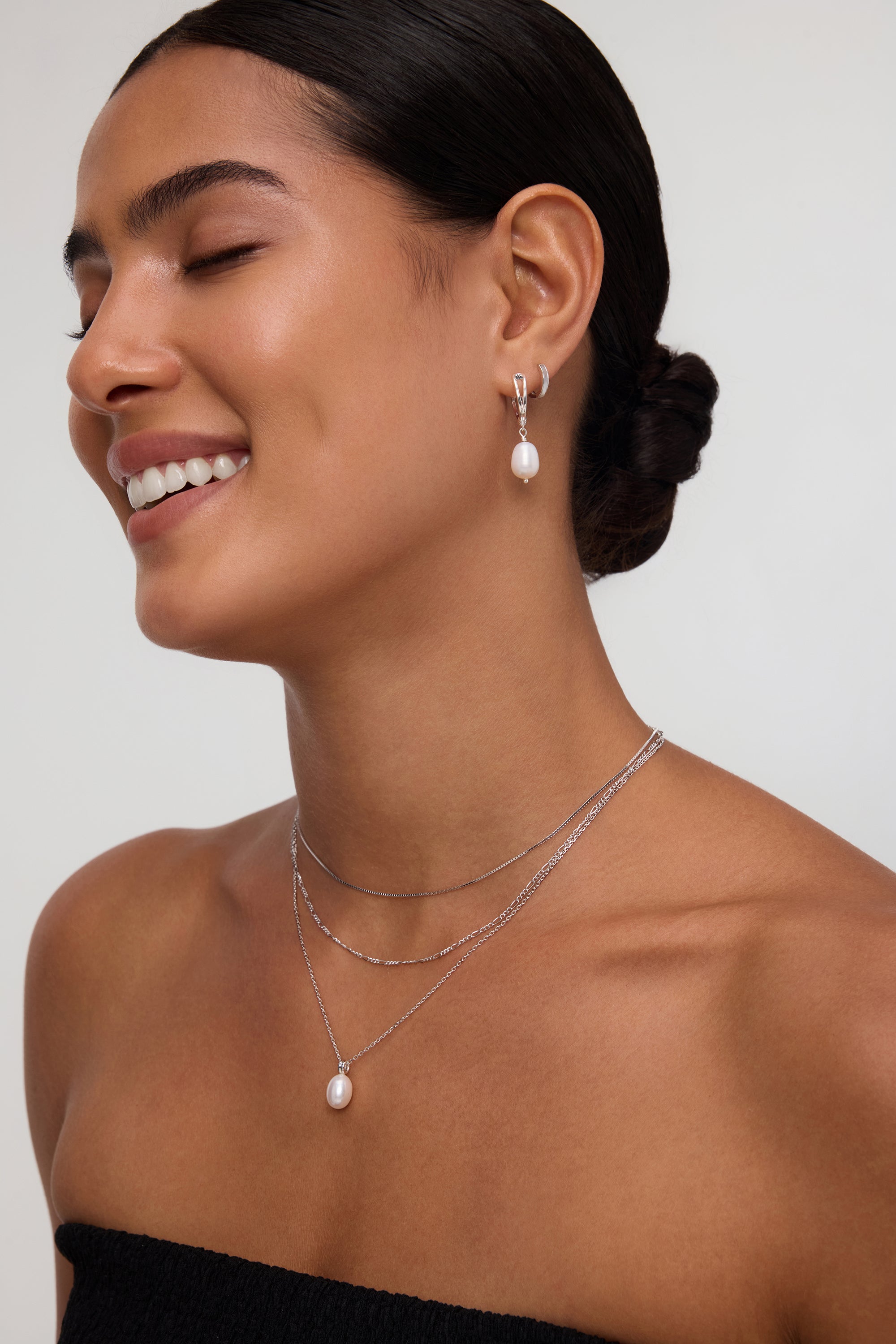 A woman with smooth, dark hair in a low bun smiles with her eyes closed, wearing a strapless black top and the Figaro And Box Chain Layered Necklace in Silver with a pearl pendant, paired with matching pearl drop earrings against a neutral background.