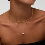 A woman wears the Mini Paperclip Double Chain Necklace in Silver with a gold chain featuring a single pearl pendant and a pearl drop earring. She is dressed in a black strapless top, her face partially visible.
