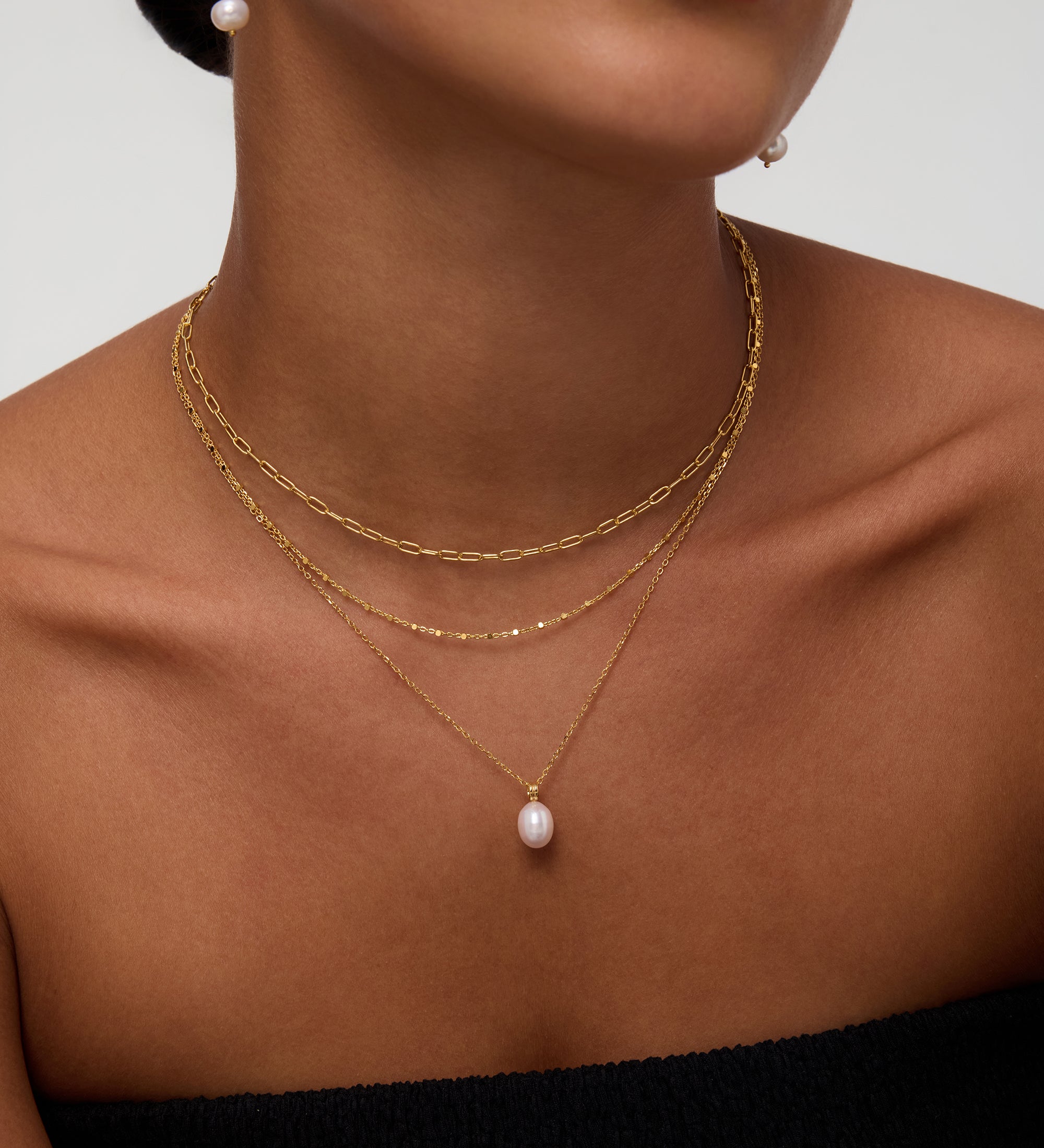 A woman wears the Mini Paperclip Double Chain Necklace in Silver with a gold chain featuring a single pearl pendant and a pearl drop earring. She is dressed in a black strapless top, her face partially visible.