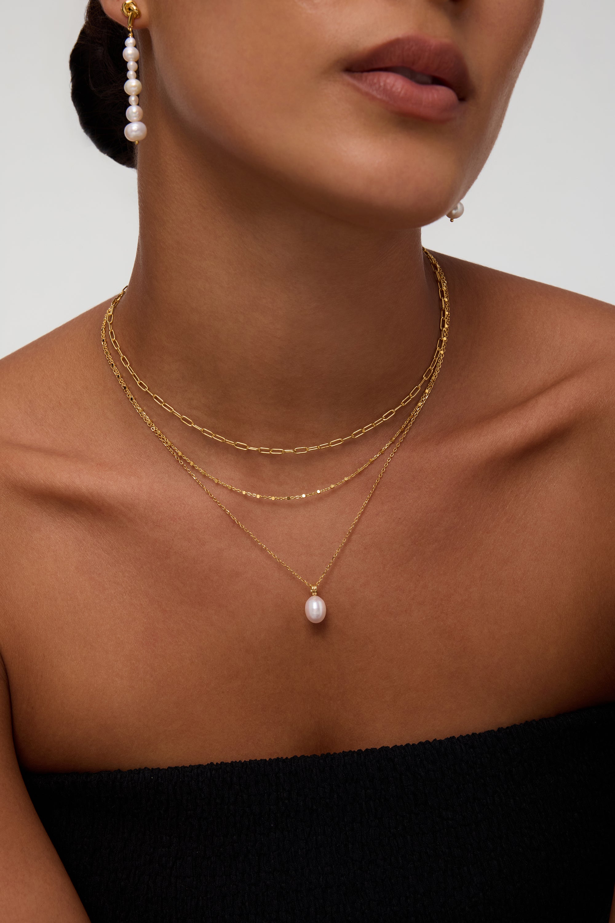 A woman wears the Mini Paperclip Double Chain Necklace in Silver with a gold chain featuring a single pearl pendant and a pearl drop earring. She is dressed in a black strapless top, her face partially visible.