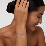 A smiling woman with dark hair in a low bun wears the Mini Paperclip Double Chain Bracelet in Silver, touching her hair and highlighting the sterling silver bracelet against her tan skin and black strapless top.