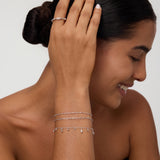 A smiling woman with dark hair in a low bun wears the Mini Paperclip Double Chain Bracelet in Silver, touching her hair and highlighting the sterling silver bracelet against her tan skin and black strapless top.
