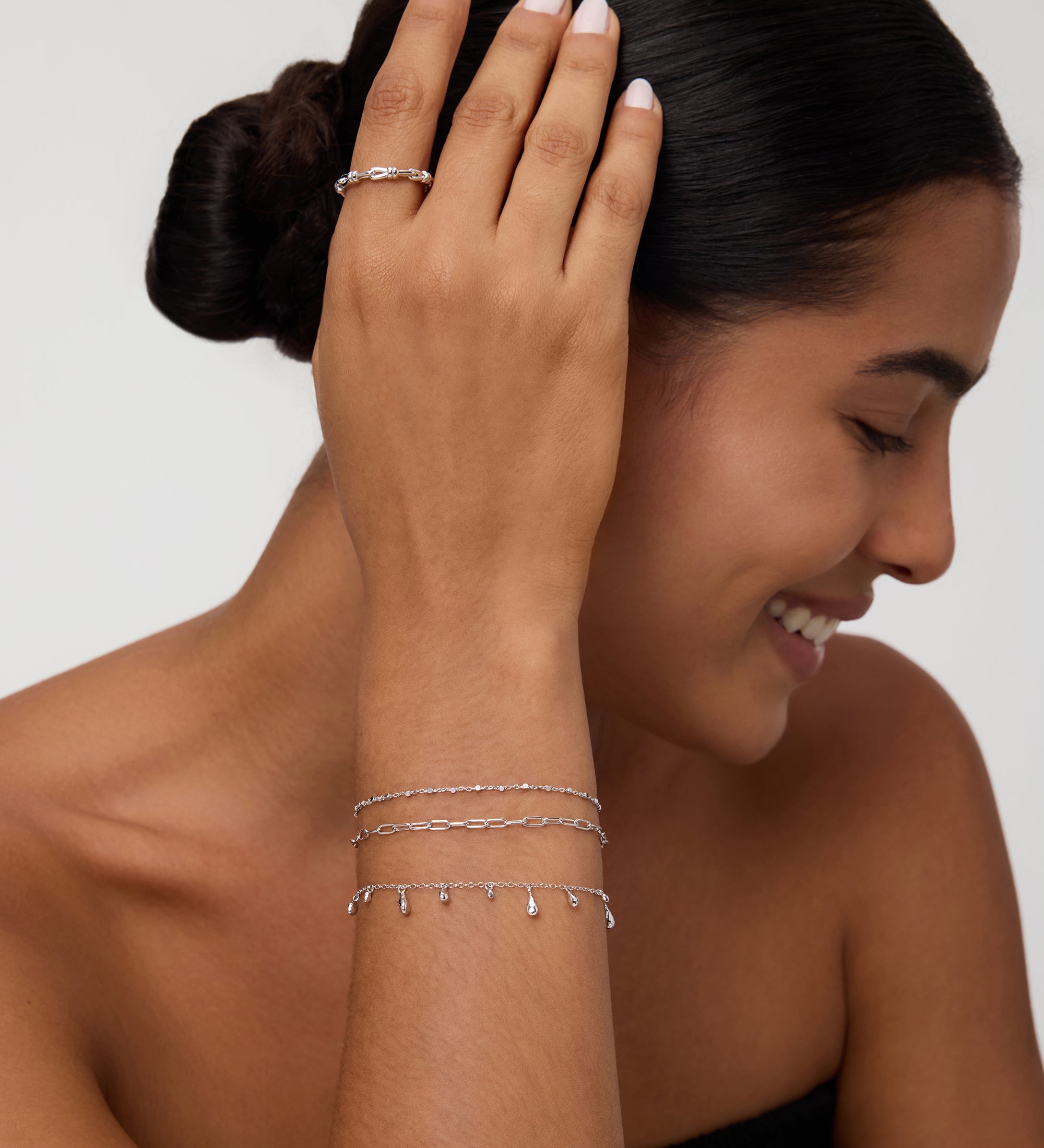A smiling woman with dark hair in a low bun wears the Mini Paperclip Double Chain Bracelet in Silver, touching her hair and highlighting the sterling silver bracelet against her tan skin and black strapless top.