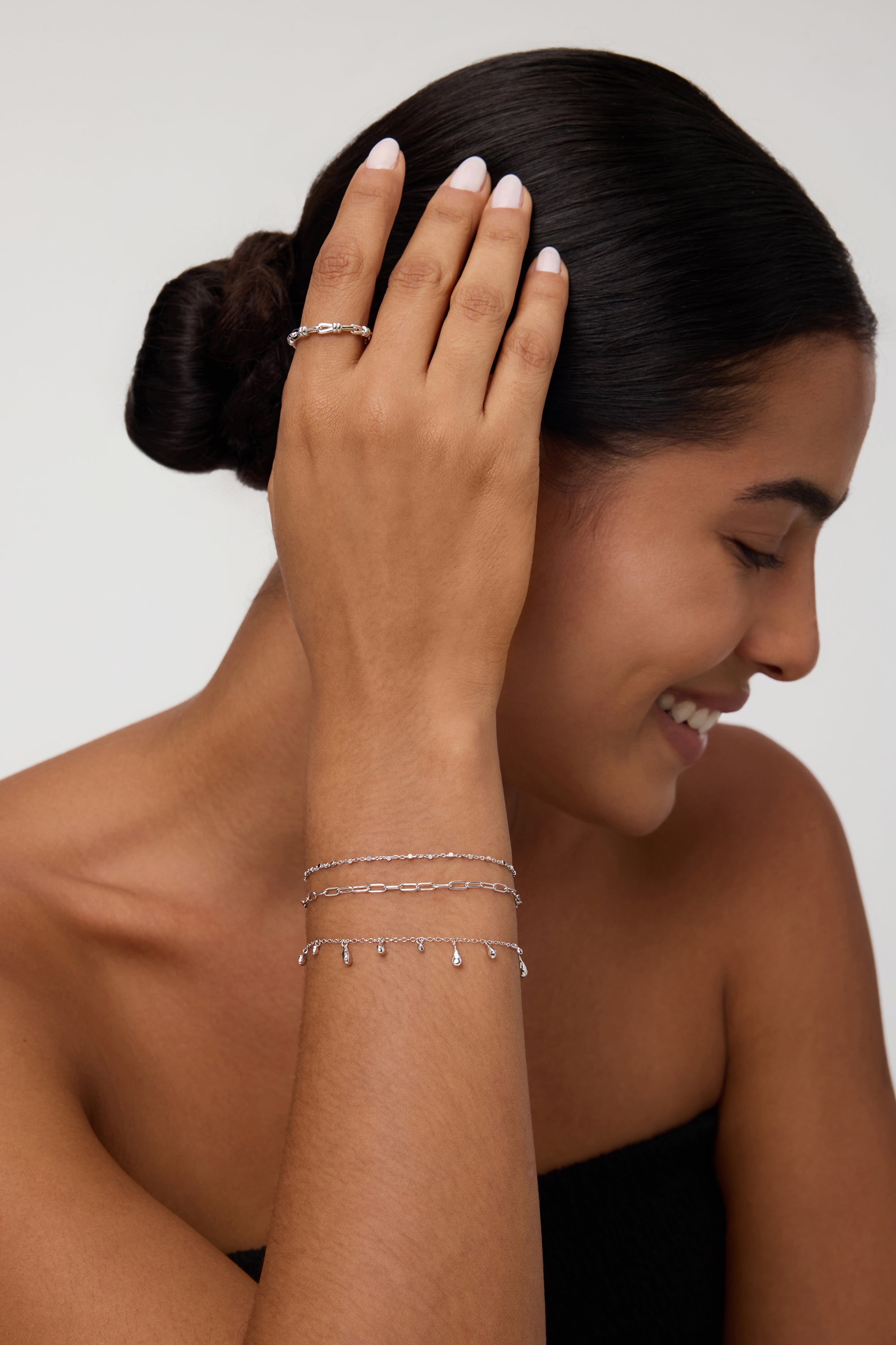 A smiling woman with dark hair in a low bun wears the Mini Paperclip Double Chain Bracelet in Silver, touching her hair and highlighting the sterling silver bracelet against her tan skin and black strapless top.