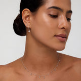 A woman with smooth, medium-toned skin and dark hair in a low bun wears the Flora Crystal Charm Huggies in Gold, featuring delicate charms, set against a neutral background.