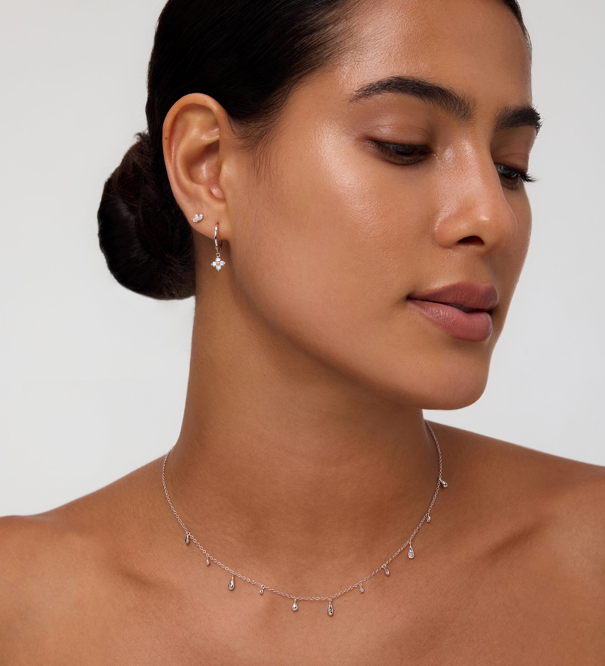 A woman with smooth, medium-toned skin and dark hair in a low bun wears the Flora Crystal Charm Huggies in Gold, featuring delicate charms, set against a neutral background.