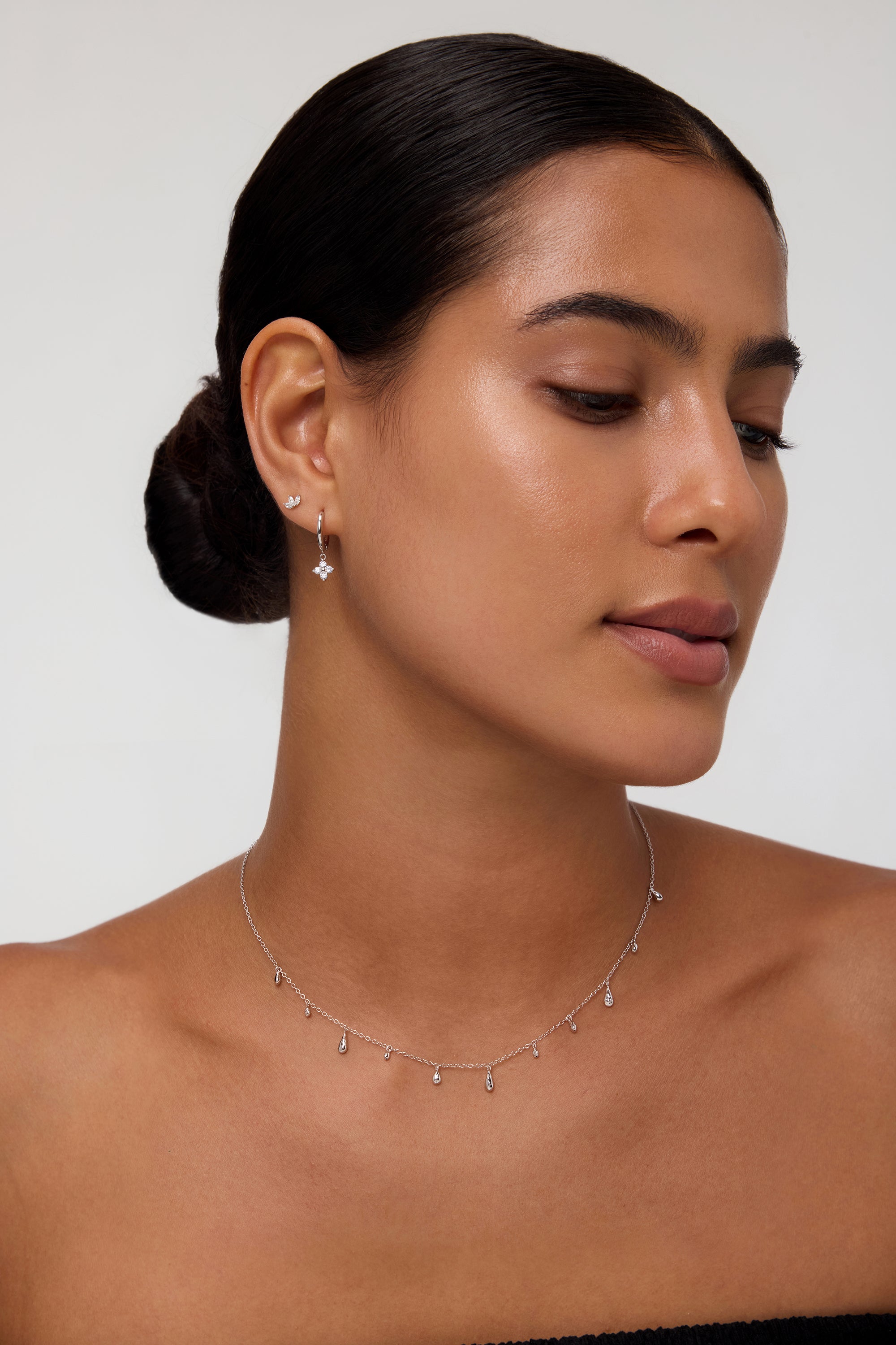 A woman with smooth, medium-toned skin and dark hair in a low bun wears the Flora Crystal Charm Huggies in Gold, featuring delicate charms, set against a neutral background.