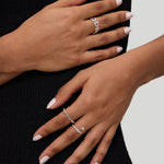 Two hands with light pink manicured nails rest on a black textured dress. Both hands wear silver rings with varying designs, including the Noa Link Ring in Gold, a delicate stacking ring that adds a modern touch to the look. The background is light gray.