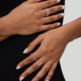 Two hands with light pink manicured nails rest on a black textured dress. Both hands wear silver rings with varying designs, including the Noa Link Ring in Gold, a delicate stacking ring that adds a modern touch to the look. The background is light gray.