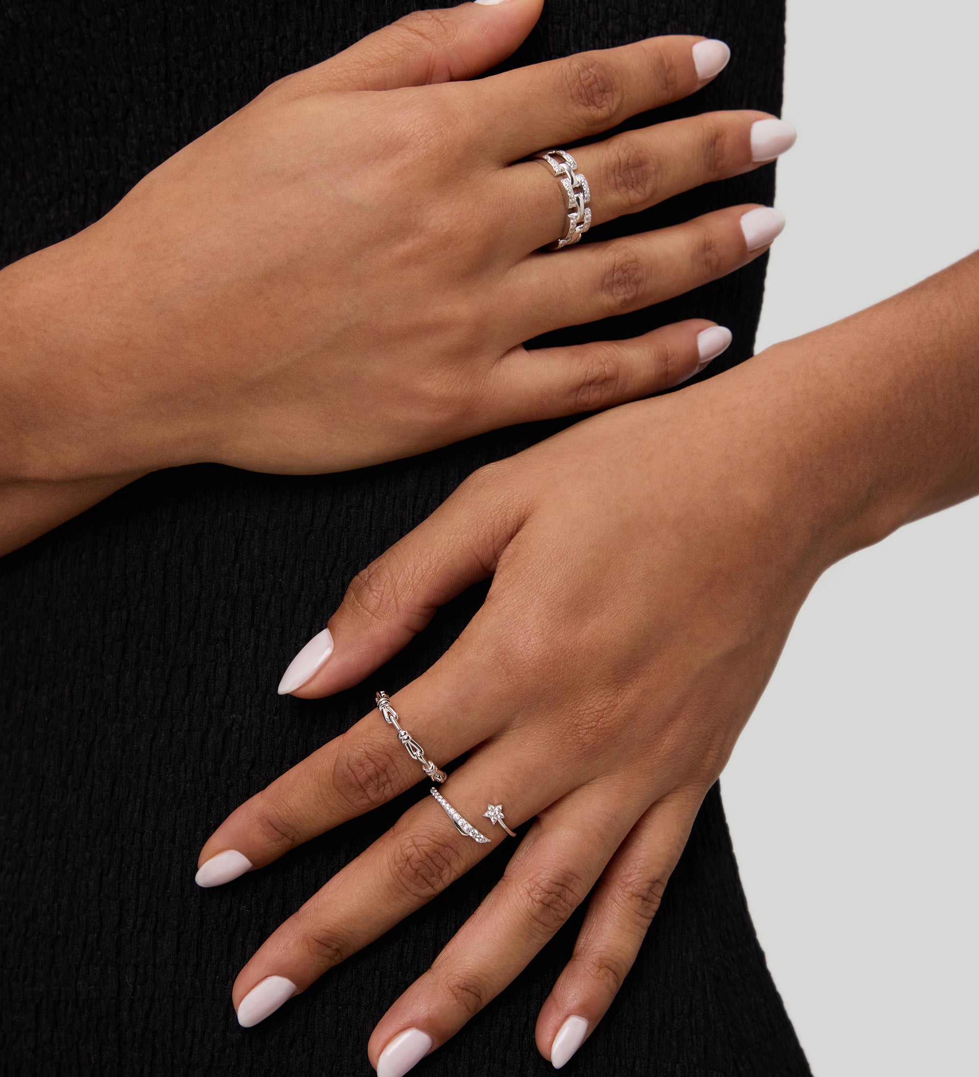 Two hands with light pink manicured nails rest on a black textured dress. Both hands wear silver rings with varying designs, including the Noa Link Ring in Gold, a delicate stacking ring that adds a modern touch to the look. The background is light gray.