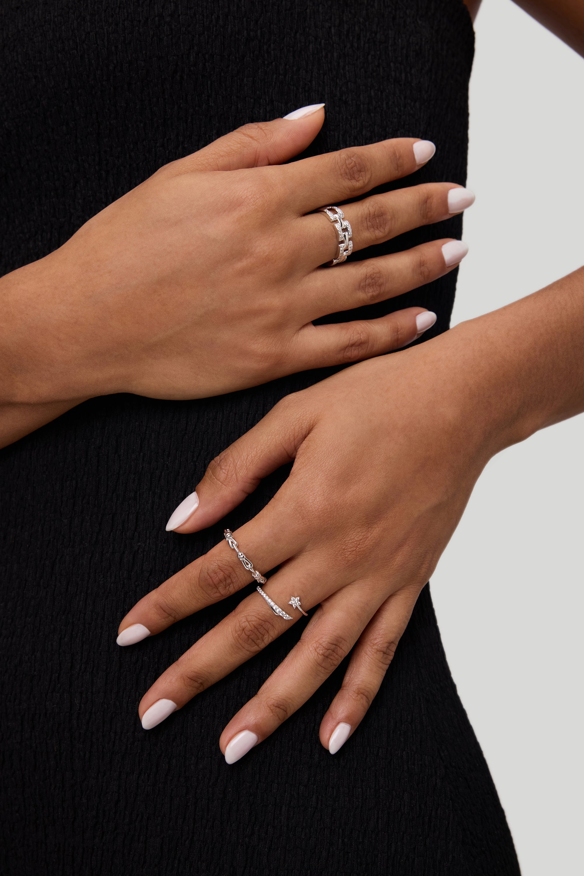 Two hands with light pink manicured nails rest on a black textured dress. Both hands wear silver rings with varying designs, including the Noa Link Ring in Gold, a delicate stacking ring that adds a modern touch to the look. The background is light gray.