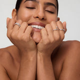 A smiling woman with eyes closed rests her chin on her hands, showcasing the Noa Link Ring in Gold among other stacking rings and silver bands on her fingers. Her nails are natural and her hair is slicked back, with a bare-shouldered, neutral background.