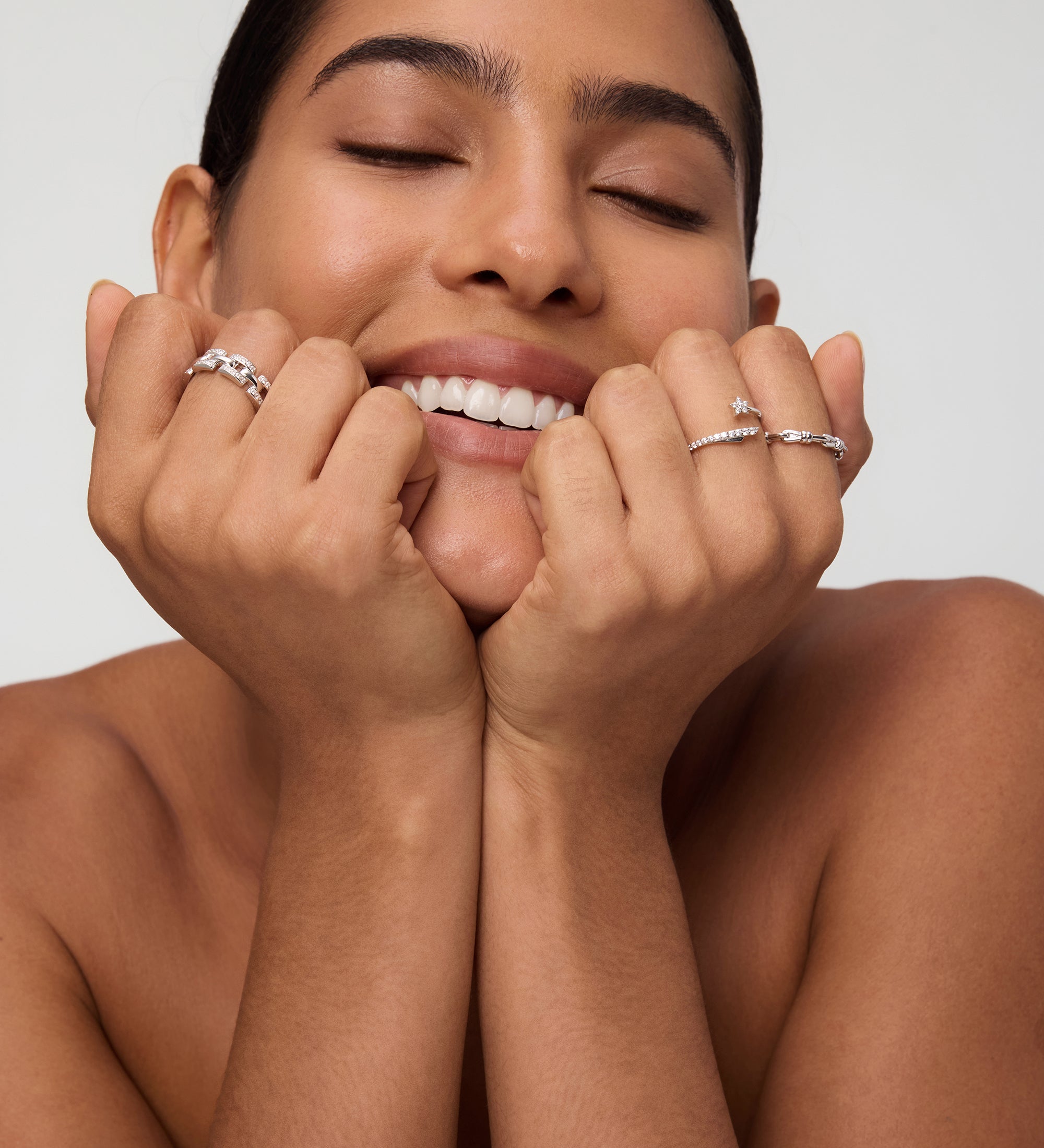 A smiling woman with eyes closed rests her chin on her hands, showcasing the Noa Link Ring in Gold among other stacking rings and silver bands on her fingers. Her nails are natural and her hair is slicked back, with a bare-shouldered, neutral background.
