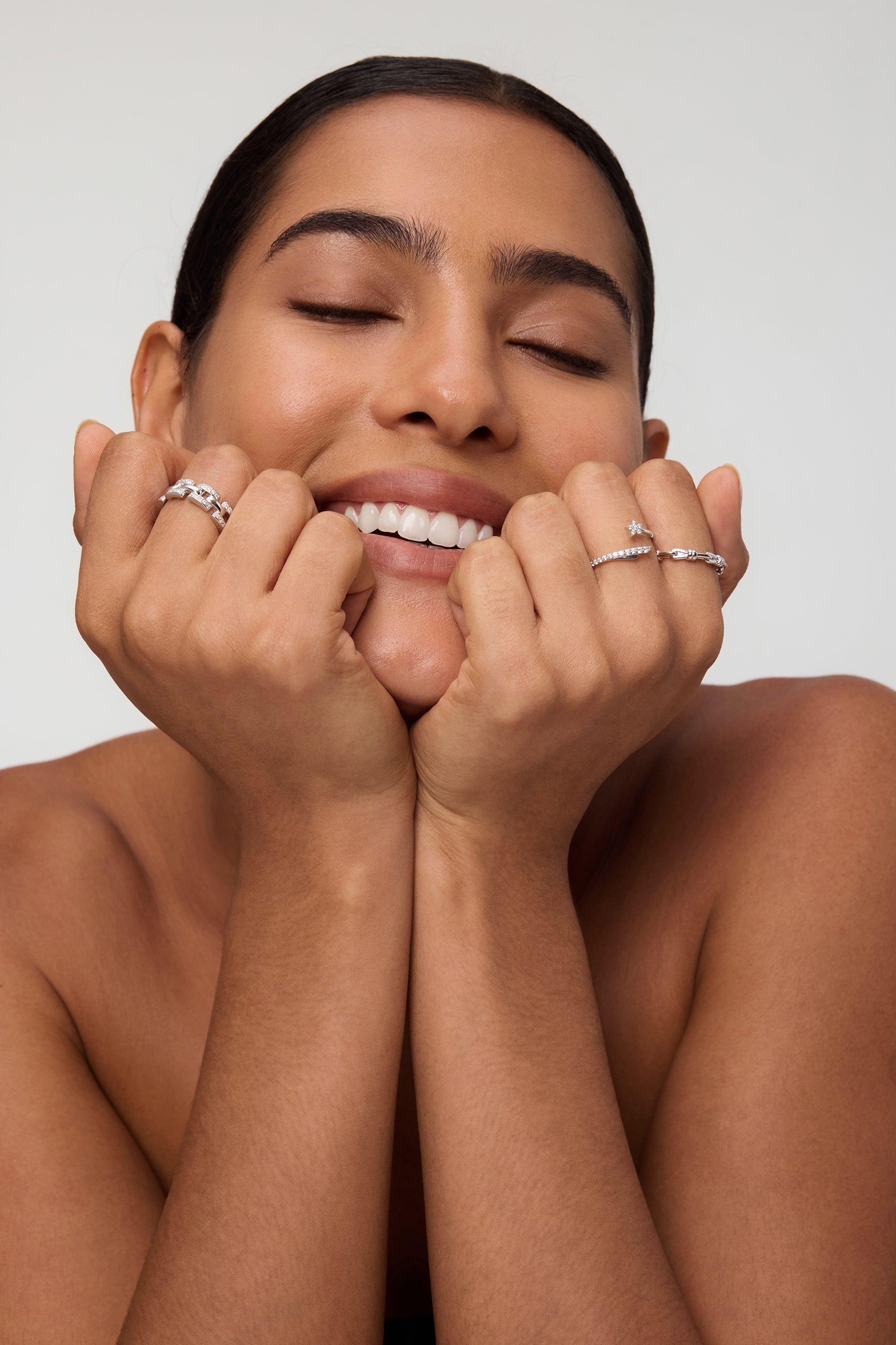 A smiling woman with eyes closed rests her chin on her hands, showcasing the Noa Link Ring in Gold among other stacking rings and silver bands on her fingers. Her nails are natural and her hair is slicked back, with a bare-shouldered, neutral background.