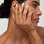 A woman with smooth skin and dark hair in a low bun touches her face, displaying the Noa Link Ring in Gold on her finger. She has natural makeup, nude-colored nails, and gazes to the side against a neutral background.